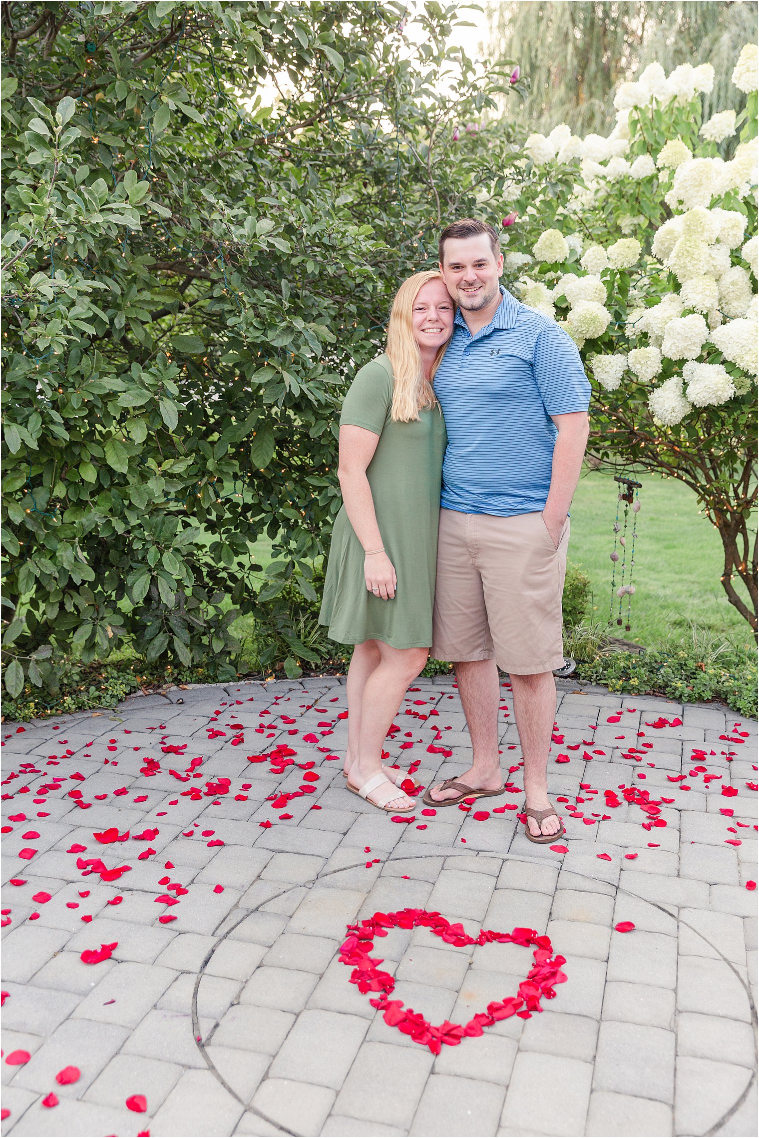 Engaged couple stand in rose petals by white flowers after surprise spring proposal in PA 