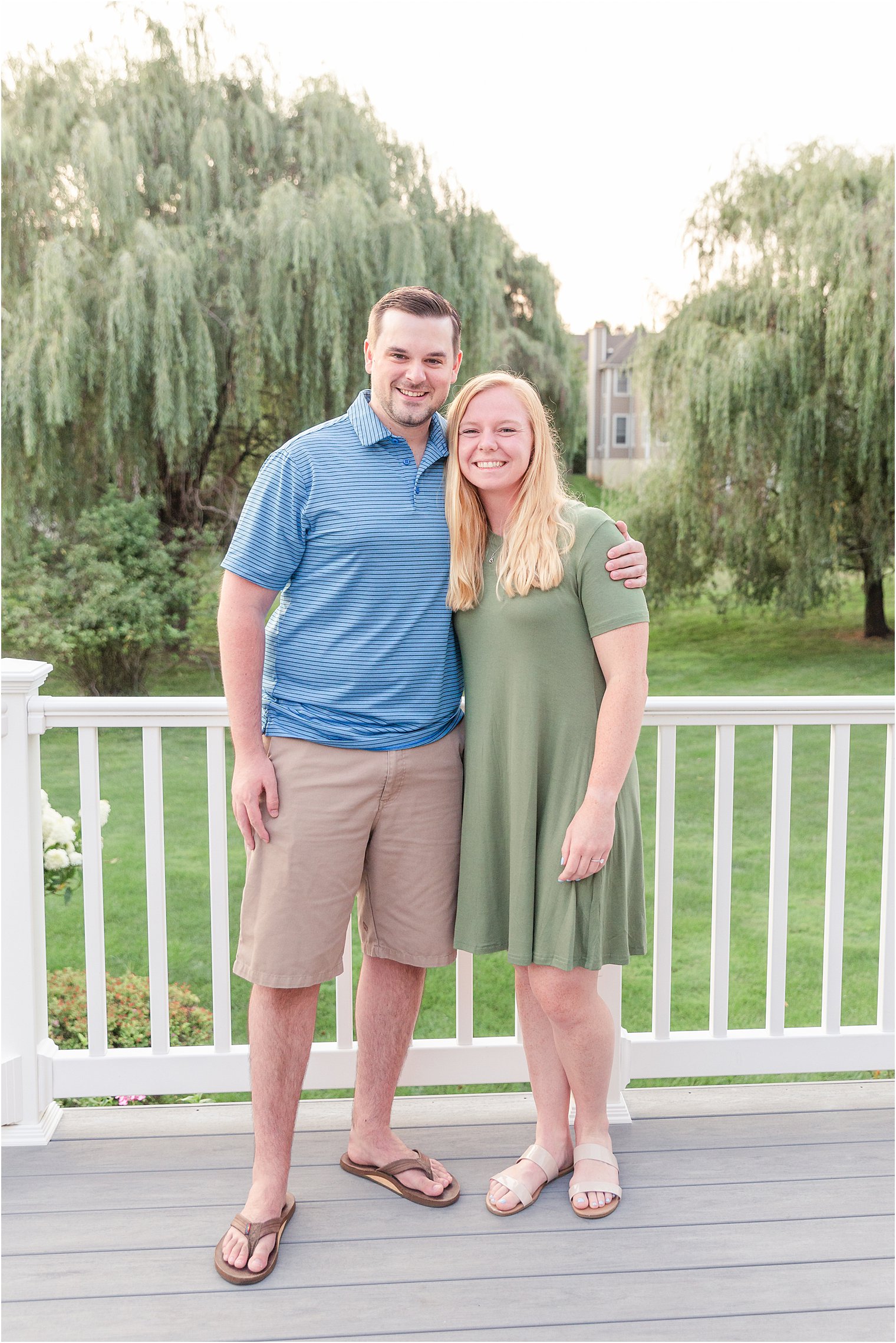 Couple stands on white deck with weeping willow trees in background after Malvern PA Backyard proposal captured by Jocelyn Cruz Photography