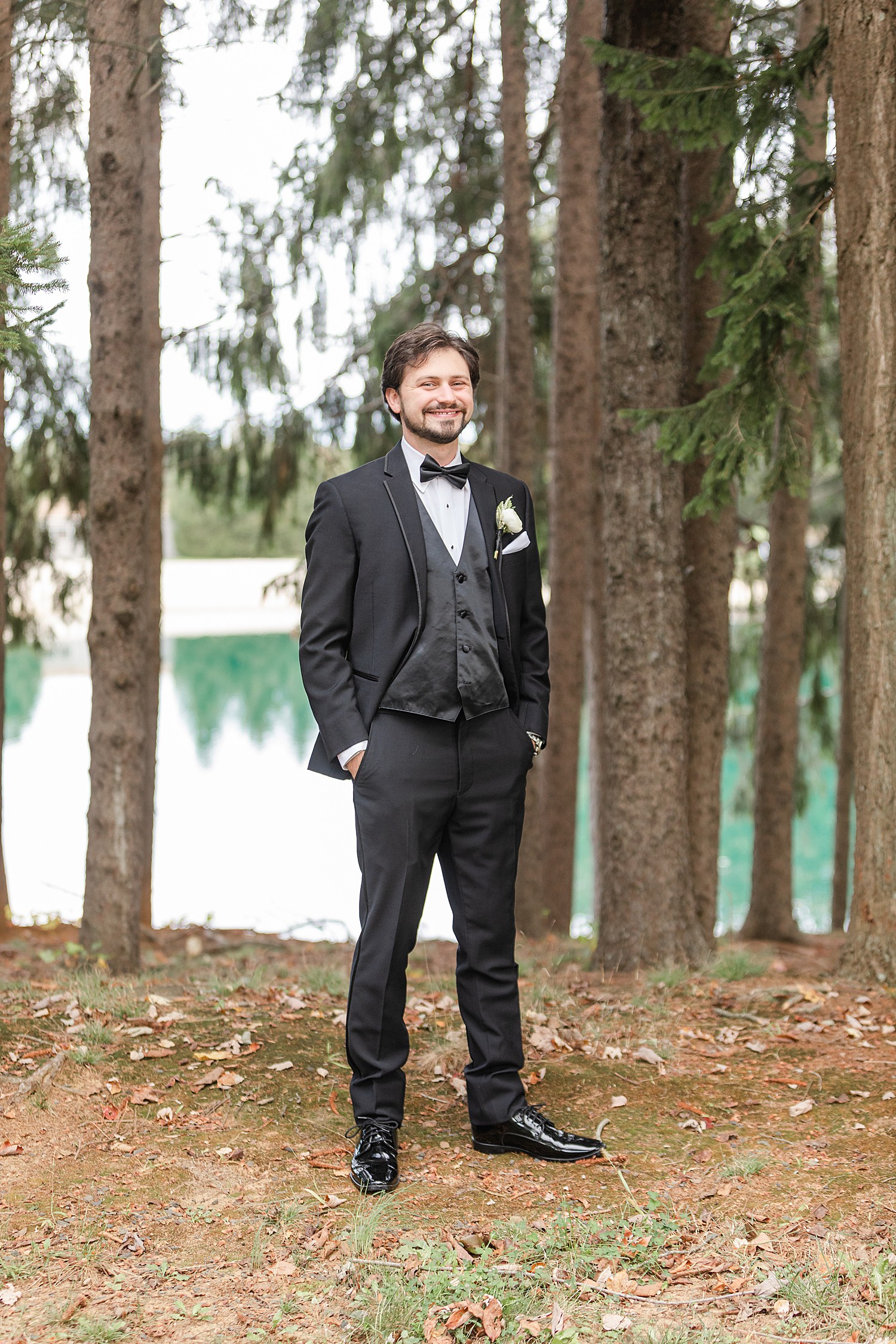 groom standing near the lake with tall pine trees in the background 