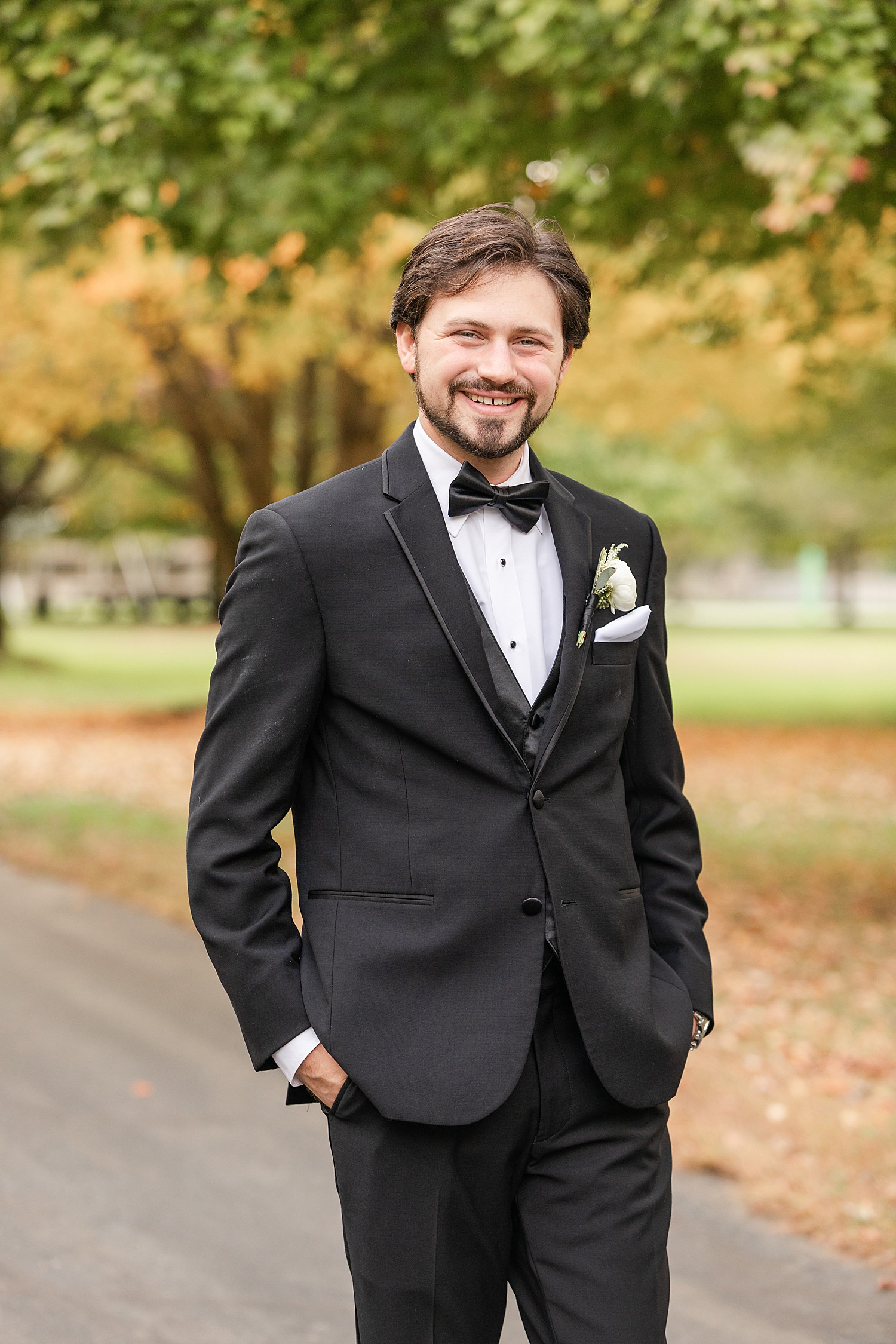 groom stands outside with beautiful fall trees in the background