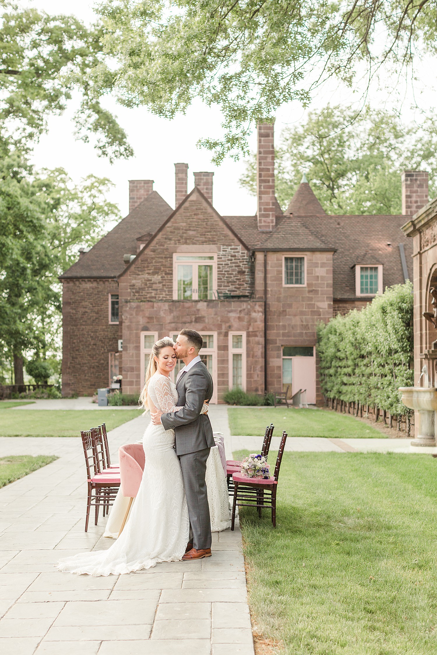 groom kisses brides head in front of old stone mansion at Tyler Gardens